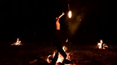 Düşük anahtar. Uzun saçlı ve çıplak gövdeli genç bir erkek, yanan bir meşaleyi alevlerin yanında siyah bir gece videosu yla açık havada döndürür. Modern fakir yanan bir personel ile hileler yapar