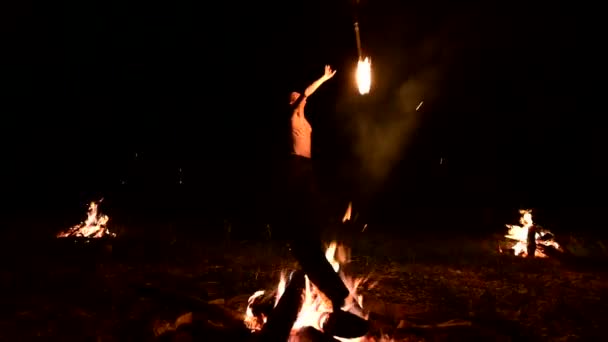 Bajo perfil. Un macho joven con el pelo largo y un torso desnudo gira una antorcha ardiente al aire libre en un video nocturno negro en cámara lenta cerca de los incendios. Fakir moderno hace trucos con un personal en llamas — Vídeo de stock