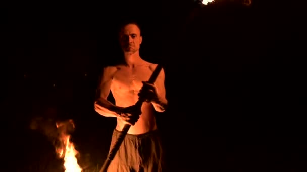 Low key. Young male with long hair and bare torso rotates burning torch outdoors on a black night video slow motion. Modern fakir does tricks with a burning staff — Stock Video