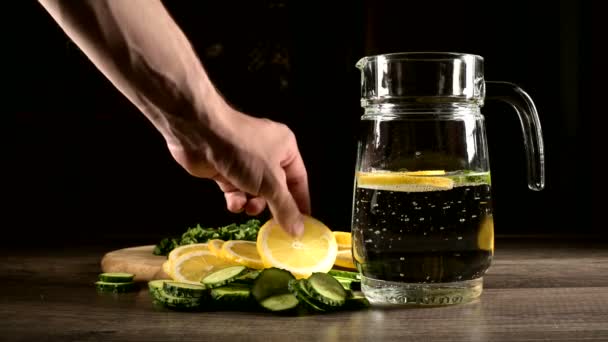 Sliced lemon cucumbers and mint leaves on a wooden cutting board next to a glass carafe with sparkling water. Male hand throwing slices of lemon in a glass of soda — Stock Video