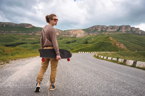 Um jovem elegante caminha ao longo de uma estrada de montanha sinuosa com um skate ou longboard em suas mãos na noite após o pôr do sol. O conceito de esportes juvenis e hobbies de viagem — Fotografia de Stock