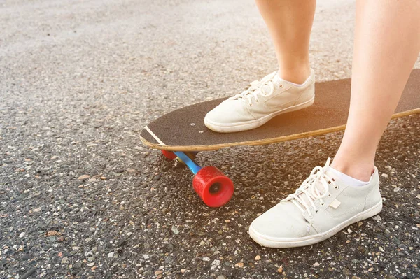 Közeli fel a női lábak rongyos cipők egy Longboard a háttérben aszfalt naplementekor. Nagy gördeszka a lány lába. Ifjúsági szabadidőkoncepció — Stock Fotó