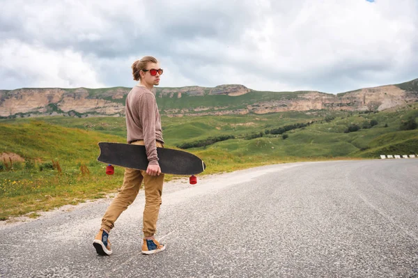 Homem jovem e elegante hipster com cabelos longos e óculos de sol andando com um longboard em suas mãos em uma estrada de asfalto rural nas montanhas no fundo de rochas épicas — Fotografia de Stock