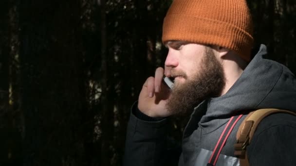 Un uomo barbuto con un cappello che parla al cellulare in una foresta di conifere. Il concetto di comunicazione cellulare in luoghi difficili da raggiungere — Video Stock