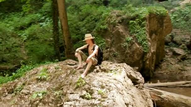 Vista aérea de un viajero masculino en un sombrero de paja y mochila se sienta en una roca contra el telón de fondo de una majestuosa cascada en el bosque entre las rocas. Viajes video 4k — Vídeo de stock