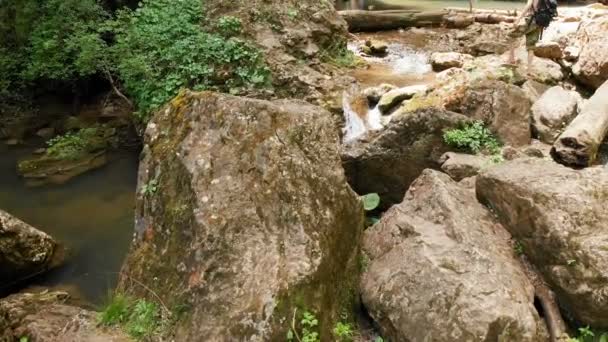 Veduta aerea di un viaggiatore di sesso maschile con un cappello di paglia e uno zaino si arrampica su una roccia sullo sfondo di una maestosa cascata nel bosco tra le rocce. Video di viaggio 4k — Video Stock