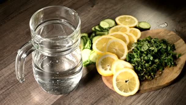 Pepinos de limón en rodajas y hojas de menta en una tabla de cortar de madera junto a una jarra de vidrio con agua con gas — Vídeos de Stock