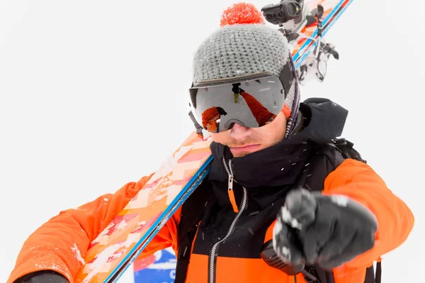 身着橙色黑色西装、肩上戴着滑雪板、在雪地里浅色背景上对着相机的专业滑雪运动员 — 图库照片