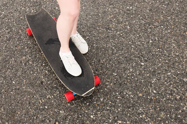 Close-up de pernas femininas em sapatilhas de pano em um longboard no fundo de asfalto ao pôr do sol. Grande skate com pernas de menina. Conceito de lazer juvenil — Fotografia de Stock