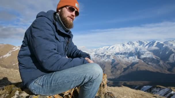 Retrato de un viajero barbudo con gafas de sol y una gorra sobre una roca sobre el telón de fondo de las montañas. Se ríe y dice — Vídeo de stock