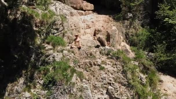 Un hombre con sombrero se sienta en el borde de un acantilado en posición de loto y medita contemplando el infinito. Vista aérea 4k — Vídeo de stock