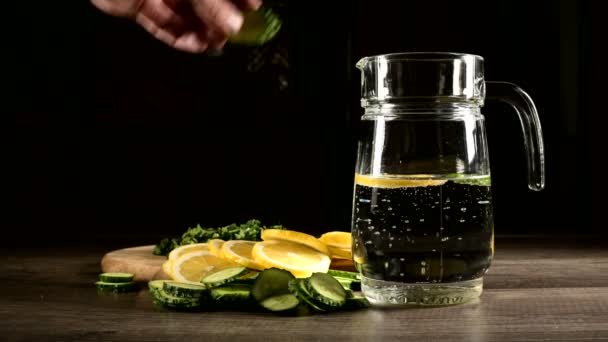 Pepinos de limón en rodajas y hojas de menta en una tabla de cortar de madera junto a una jarra de vidrio con agua con gas. Macho mano lanzando rodajas de pepino en un vaso de refresco — Vídeos de Stock