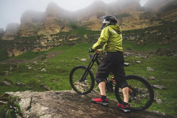 Vista lateral de un hombre en una bicicleta de montaña de pie en un terreno rocoso y mirando una roca. El concepto de una bicicleta de montaña y mtb cuesta abajo —  Fotos de Stock