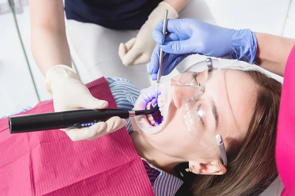 Κοντινό πλαίσιο ενός οδοντιάτρου και νοσοκόμου που φτιάχνει μια αποξηραμένη νέα γέμιση με υπεριώδης νεαρή γυναίκα ασθενή στο οδοντιατρείο — Φωτογραφία Αρχείου