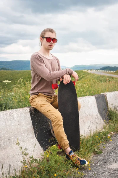 Homem elegante jovem com cabelos longos em óculos de sol está sentado em um chipper com um longboard em suas mãos em uma estrada de asfalto país no fundo de rochas e nuvens — Fotografia de Stock