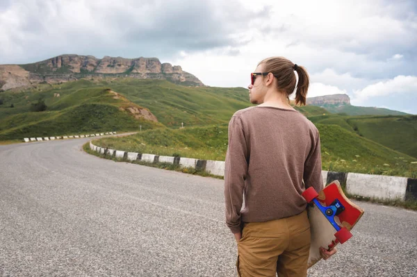 Joven hombre elegante con el pelo largo reunido en una cola de caballo y en gafas de sol se encuentra con un longboard en sus manos en un camino de asfalto país en las montañas en el fondo de rocas épicas — Foto de Stock