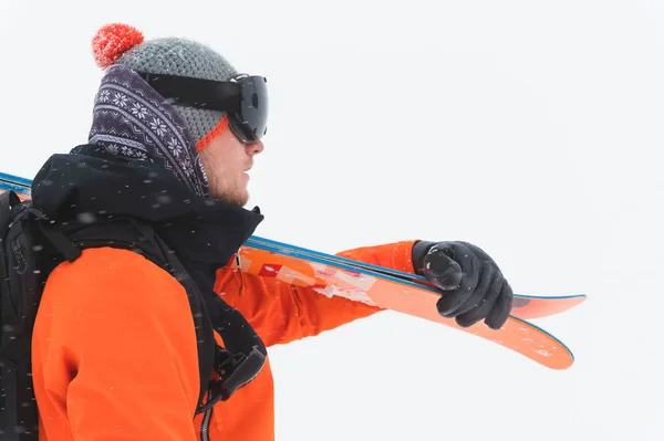 Retrato de un atleta profesional esquiador en una chaqueta naranja con una máscara negra y con esquís en el hombro se ve a un lado. Fondo gris durante tormenta de nieve — Foto de Stock