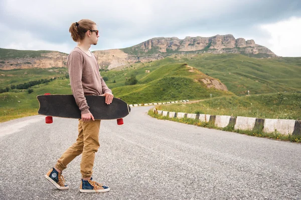 Şık bir genç adam gün batımından sonra akşam elinde bir paten veya longboard ile dolambaçlı bir dağ yolu boyunca yürüyor. Gençlik spor ve seyahat hobileri kavramı