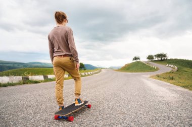 Uzun saçlı genç şık adam bir at kuyruğu ve güneş gözlüğü toplanan epik kayalar arka planda dağlarda bir ülke asfalt yol üzerinde bir Longboard ile duruyor