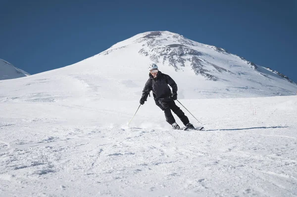 A bearded mature aged male skier in a black ski suit descends along the snowy slope of a ski resort amid two peaks of Mount Elbrus. The concept of sports in adulthood