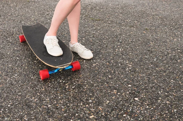Close-up de pernas femininas em sapatilhas de pano em um longboard no fundo de asfalto ao pôr do sol. Grande skate com pernas de menina. Conceito de lazer juvenil — Fotografia de Stock