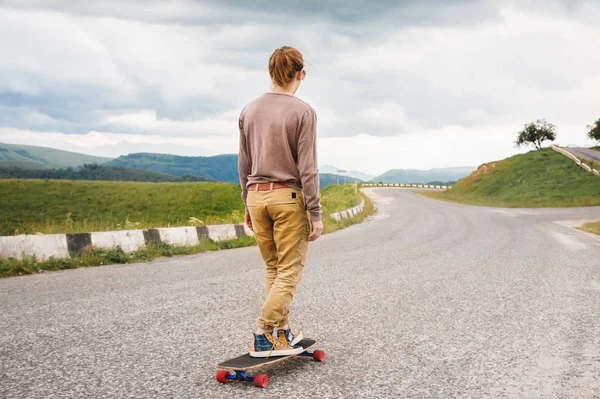 Стильний чоловік їде на дошці на скейтборді вздовж асфальтової заміської дороги ввечері проти неба гір і пагорбів — стокове фото
