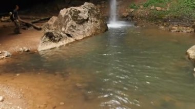 Sırt çantası olan bir erkek gezginin havadan görünümü, kayalıkların arasındaki ormanda görkemli bir şelalenin önündeki uçuruma tırmanıyor. Seyahat video 4k