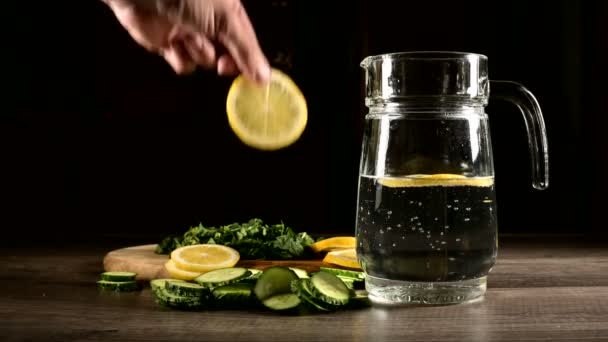 Sliced lemon cucumbers and mint leaves on a wooden cutting board next to a glass carafe with sparkling water. Male hand throwing slices of lemon in a glass of soda — Stock Video