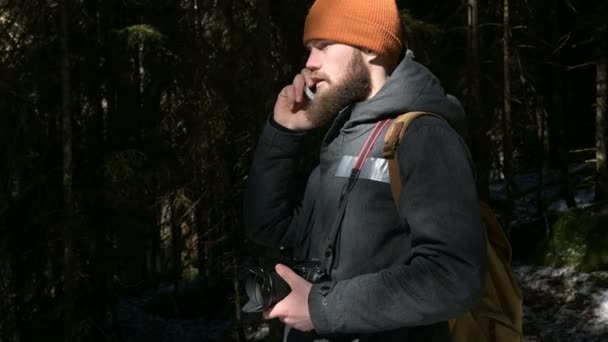Ein bärtiger Mann mit Hut, der in einem Nadelwald mit einem Handy telefoniert. das Konzept der zellularen Kommunikation an schwer zugänglichen Orten — Stockvideo