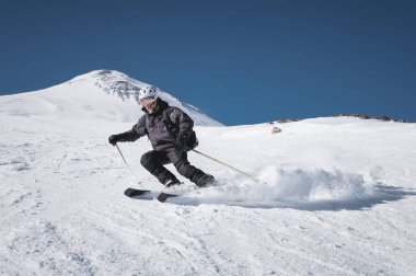 Siyah kayak elbisesi giymiş sakallı, yaşlı ve erkek kayakçı, Elbrus Dağı'nın iki zirvesinin ortasında bir kayak merkezinin karlı yamacıboyunca iner. Yetişkinlikte spor kavramı