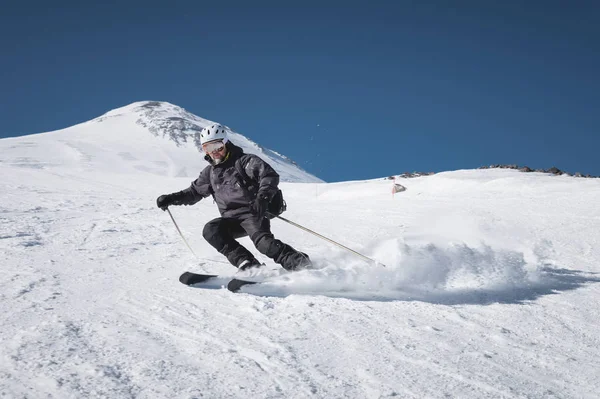 Un esquiador macho maduro barbudo con un traje de esquí negro desciende a lo largo de la ladera nevada de una estación de esquí en medio de dos picos del Monte Elbrus. El concepto de deporte en la edad adulta —  Fotos de Stock