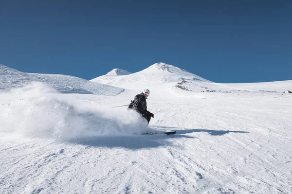 Un esquiador macho maduro barbudo con un traje de esquí negro desciende a lo largo de la ladera nevada de una estación de esquí en medio de dos picos del Monte Elbrus. El concepto de deporte en la edad adulta — Foto de Stock