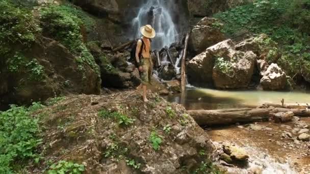 Veduta aerea di un viaggiatore di sesso maschile con un cappello di paglia e uno zaino si arrampica su una roccia sullo sfondo di una maestosa cascata nel bosco tra le rocce. Video di viaggio 4k — Video Stock