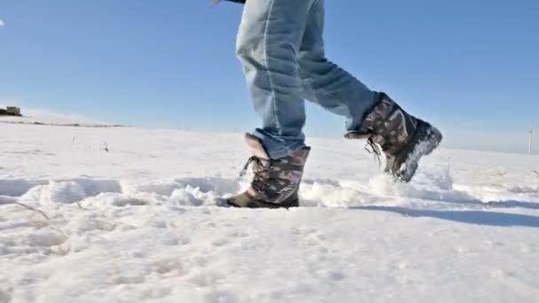 冬の晴れた日に雪の中を歩くジーンズと暖かい靴の男性の足に続きます。クローズアップローアングル。側面図 — ストック動画