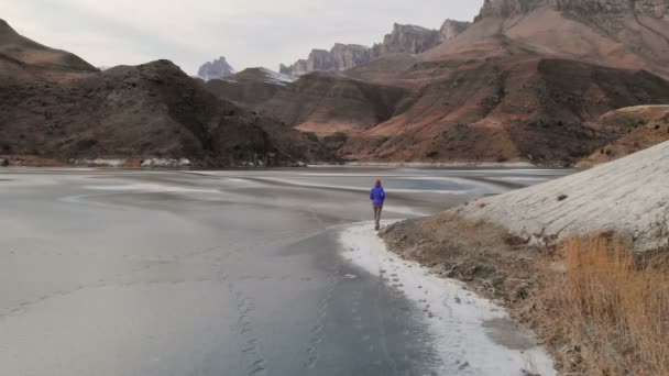 冬季黄昏时分,一位女摄影师在史诗般的岩石和山脉背景下沿着冰冻的山湖岸边行走的鸟瞰图. — 图库视频影像