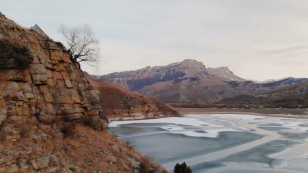 Vista aérea de uma encosta de montanha e um penhasco na margem de um lago de montanha congelado contra o pano de fundo de rochas e montanhas épicas após o pôr do sol ao anoitecer no inverno. Tecla baixa — Vídeo de Stock