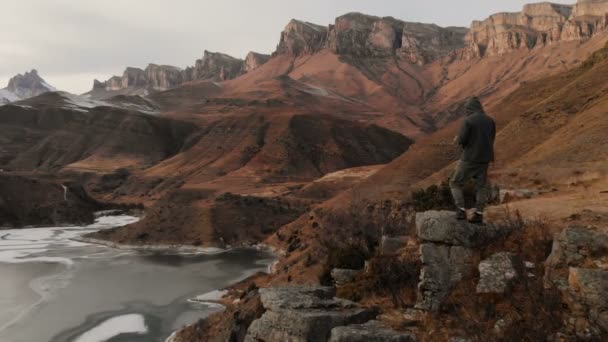 Uma vista aérea de um homem em um capuz está na borda de um penhasco perto de um lago da montanha contra o pano de fundo de rochas e montanhas épicas após o pôr do sol no crepúsculo no inverno . — Vídeo de Stock