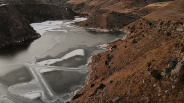 Luftaufnahme eines Berghanges und einer Klippe am Ufer eines gefrorenen Bergsees vor der Kulisse epischer Felsen und Berge nach Sonnenuntergang in der Abenddämmerung im Winter. leise — Stockvideo