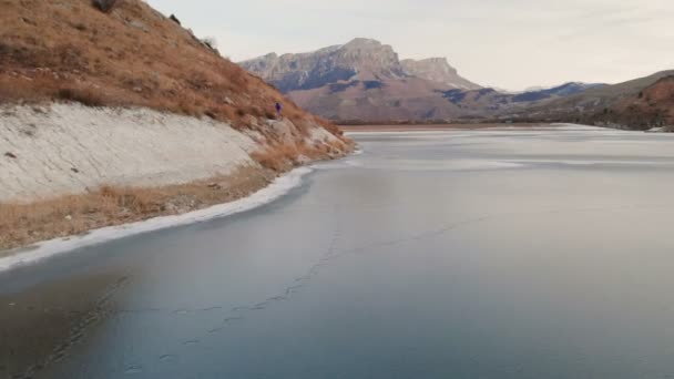 Aerial view of a woman photographer walking along the shore of a frozen mountain lake against the backdrop of epic rocks and mountains after sunset at dusk in winter. — Stock Video