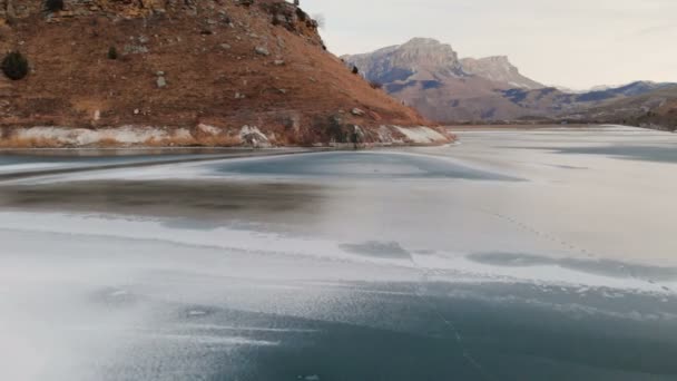 冬季黄昏时分,一位女摄影师在史诗般的岩石和山脉背景下沿着冰冻的山湖岸边行走的鸟瞰图. — 图库视频影像