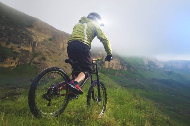 Kask ve maskeli yaşlı erkek atlet, bulutlu bir günde yayla kayalarının ve alçak bulutların arka planına karşı çimenli bir yamaçta bir dağ bisikletine biner. Yokuş aşağı dağ bisikleti konsepti
