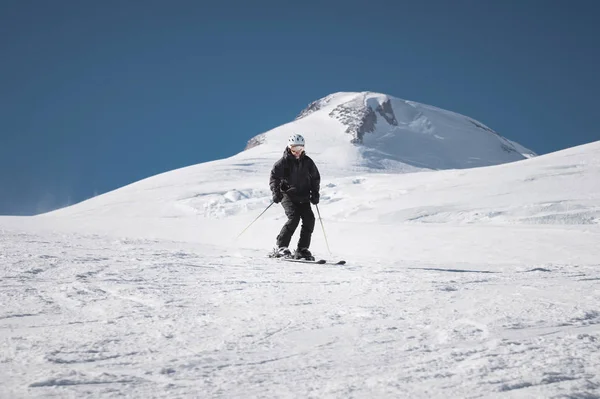 Lungo il pendio innevato di un comprensorio sciistico in mezzo a due cime del Monte Elbrus scende uno sciatore di sesso maschile con barba matura e tuta nera. Il concetto di sport in età adulta — Foto Stock
