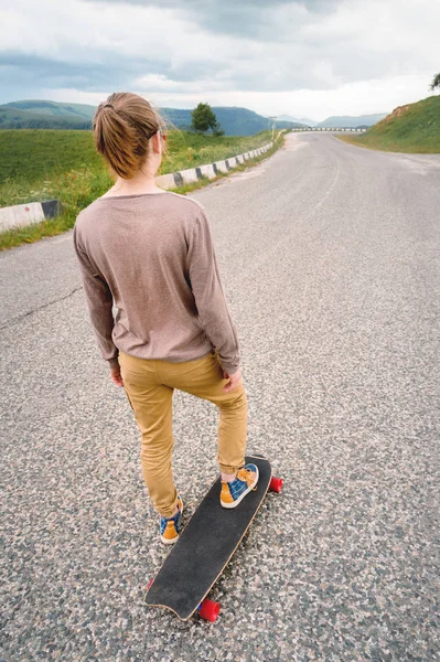 Young stylish man with long hair gathered in a ponytail and in sunglasses stands with a longboard on a country asphalt road in the mountains on the background of epic rocks — Stock Photo, Image