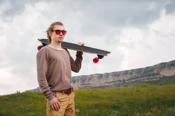 Jovem elegante de pé ao longo de uma estrada de montanha sinuosa com um skate ou longboard em seu ombro à noite após o pôr do sol. O conceito de esportes juvenis e hobbies de viagem — Fotografia de Stock