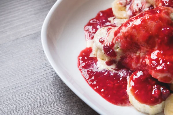 Closeup Vanilla ice cream poured with strawberry or raspberry jam with sliced banana slices on a white plate on a dark wooden table. You can do this at home on vacation