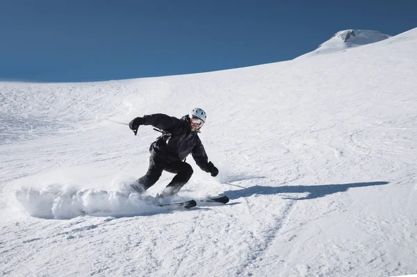 Brodaty dojrzały mężczyzna narciarz w czarnym garnitur narciarski schodzi wzdłuż ośnieżone stoku ośrodka narciarskiego pośród dwóch szczytów góry Elbrus. Koncepcja sportu w dorosłości — Zdjęcie stockowe
