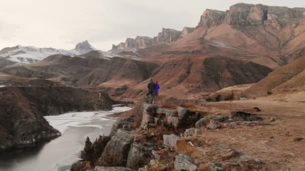 Luchtfoto van een jong stel staande op de rand van een klif in de buurt van een bergmeer tegen de achtergrond van epische rotsen en bergen na zonsondergang in de schemering. — Stockvideo