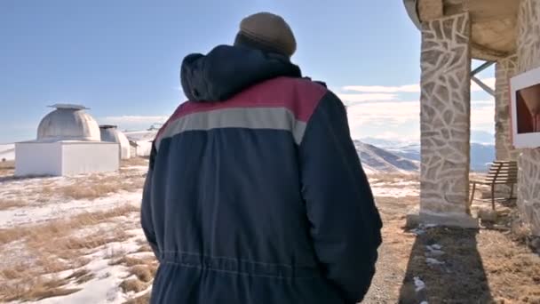 Seguindo um pesquisador masculino em jeans e um uniforme quente caminhando pela neve em um dia ensolarado de inverno entre os edifícios científicos do observatório solar. Estação de investigação científica — Vídeo de Stock