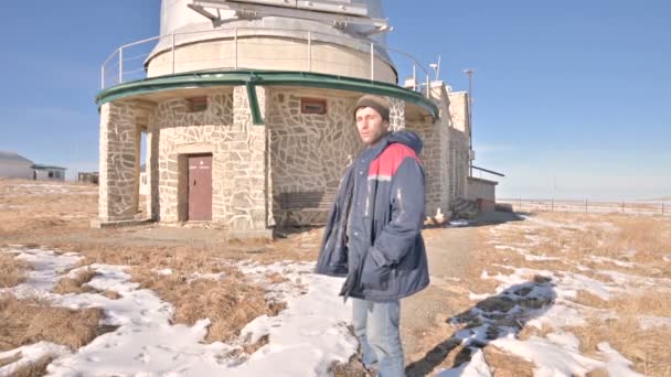 Mužský badatel v džínách a teplá uniforma v zimním slunném dnu proti pozadí vědeckých staveb u sluneční observatoře. Vědecká výzkumná stanice v kavkazských horách — Stock video