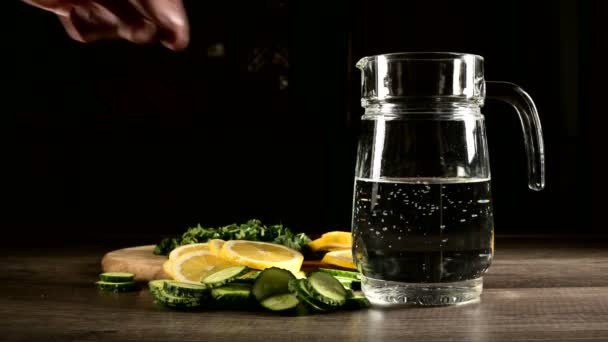 Zitronengurken und Minzblätter in Scheiben geschnitten auf einem Holzschneidebrett neben einer Glaskaraffe mit sprudelndem Wasser. Männliche Hand wirft Zitronenscheiben in ein Glas Limo — Stockvideo